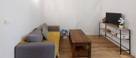 A small, cozy room with a grey sofa, a wooden table, and a TV on a shelf.