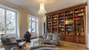 A spacious living room with a large bookshelf, comfortable seating, and a view through large windows.