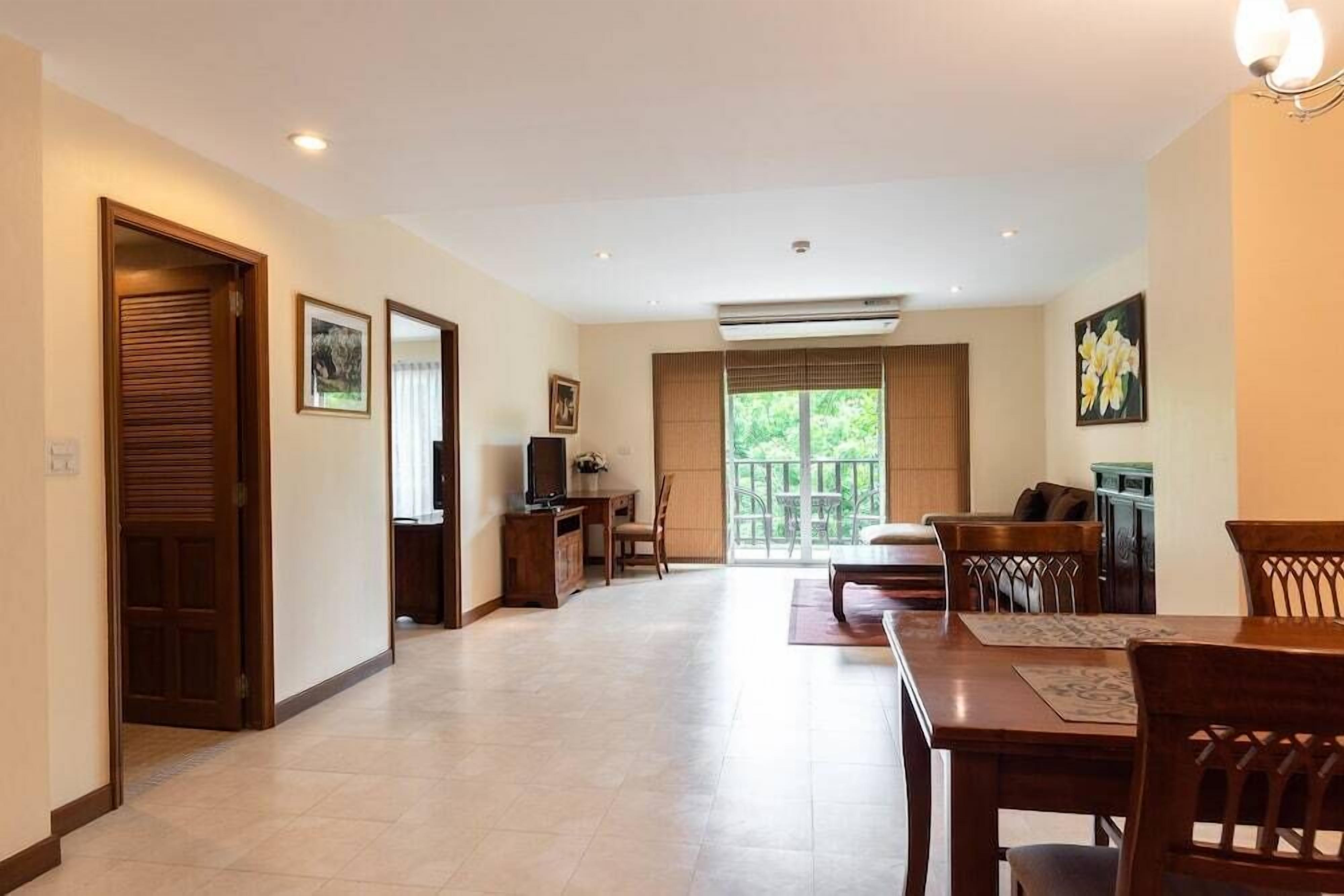 A spacious living area with a dining table, chairs, a TV, and a balcony with a view of greenery.