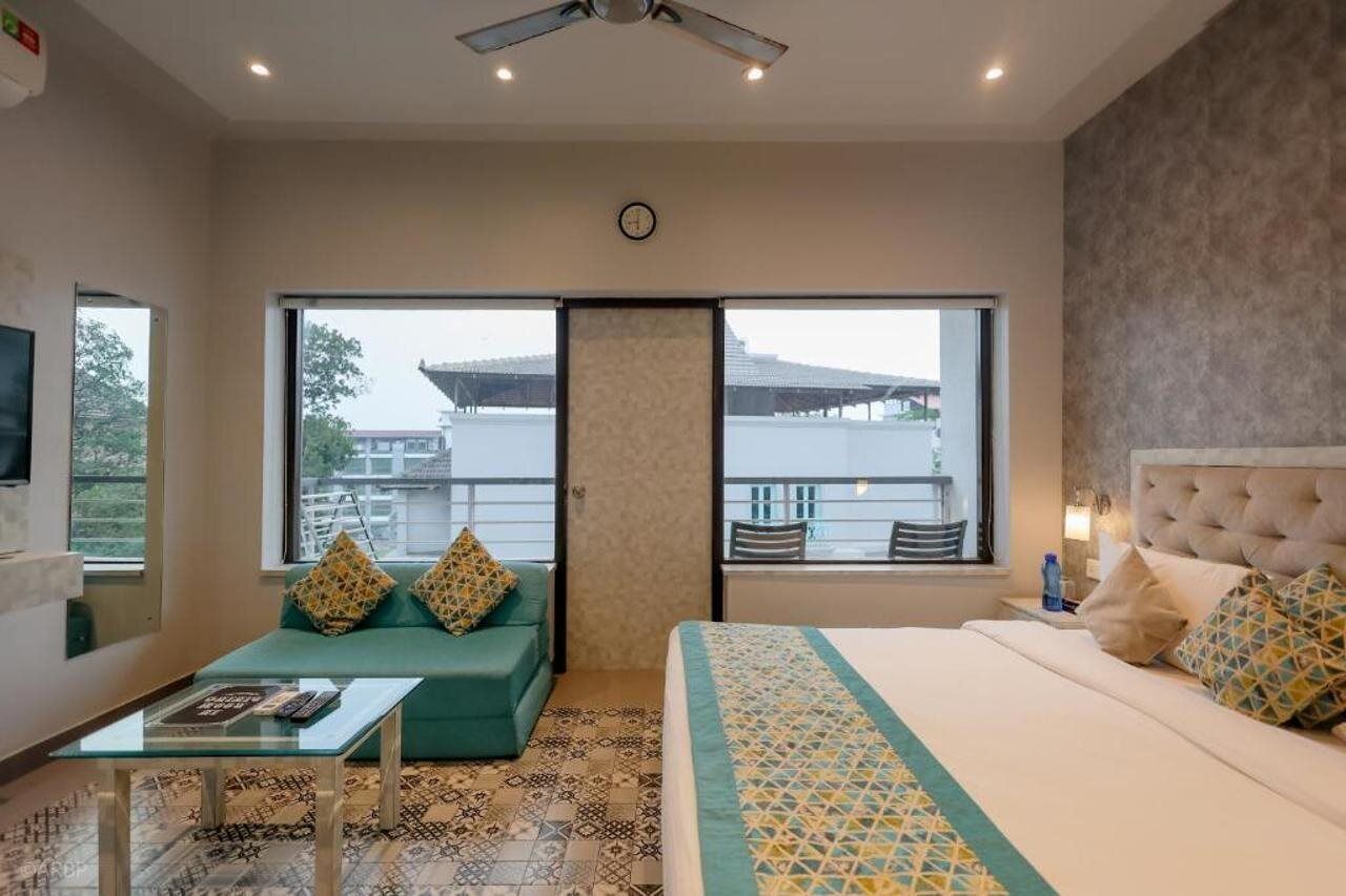 A hotel room with a bed, a sofa, a small table, and a balcony with a view of buildings.