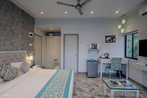 A hotel room with a bed, a desk, a chair, a small fridge, a TV, and a window. - La Sunila Suites (Arpora)