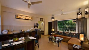 A modern living room with a sofa, coffee table, and dining area. There is a large window with curtains, a ceiling fan, and wall-mounted artwork. - La Sunila Suites (Arpora)