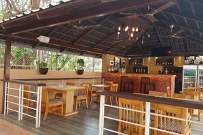 An outdoor bar area with wooden tables and chairs, a counter with a colorful patterned tablecloth, and a ceiling fan. - La Sunila Suites (Arpora)