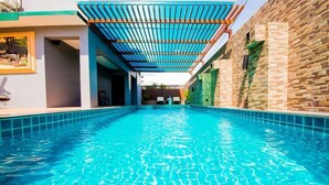 Ein Pool mit einer gefliesten Kante, ein überdachter Bereich mit blauem Dach und eine Backsteinmauer an einer Seite.