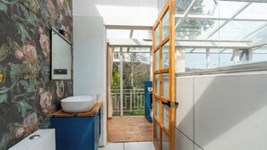 A modern bathroom with a white sink, blue cabinet, and a wooden door leading to a balcony with a view of trees and a building.