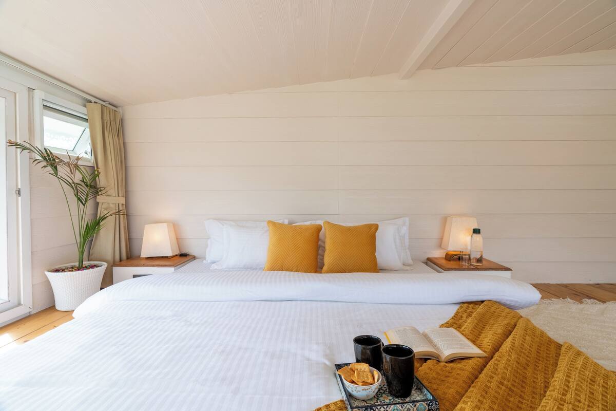 A neatly made bed with white bedding and yellow pillows, a bedside table with a lamp, a potted plant, and a tray with snacks and a book.
