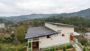A white building with a green roof and a balcony, surrounded by a mountainous landscape.