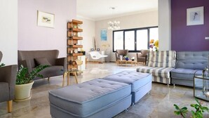 A modern living room with a grey sofa, a striped sofa, a glass coffee table, and a grey ottoman. There is a wooden shelf unit against the wall and a potted plant on the floor.