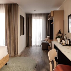 A hotel room with a bed, a desk, a chair, a sofa, and a large window with curtains. - Strand Suites By Neu Collective (Gzira)