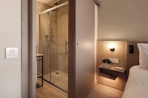A modern hotel room with a glass-enclosed shower, a bedside table with a plant, and a bedside lamp. - Strand Suites By Neu Collective (Gzira)