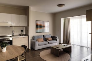 A modern living space with a kitchen, a grey sofa with cushions, a coffee table, and a dining area with a round table and chairs. - Strand Suites By Neu Collective (Gzira)