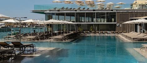 Zona de piscina con tumbonas y sombrillas, una piscina azul cristalina y un edificio moderno con grandes ventanales.