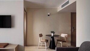 A modern living room with a round black table, two wooden chairs, a grey sofa, a TV mounted on the wall, and a door. - Strand Suites By Neu Collective (Gzira)