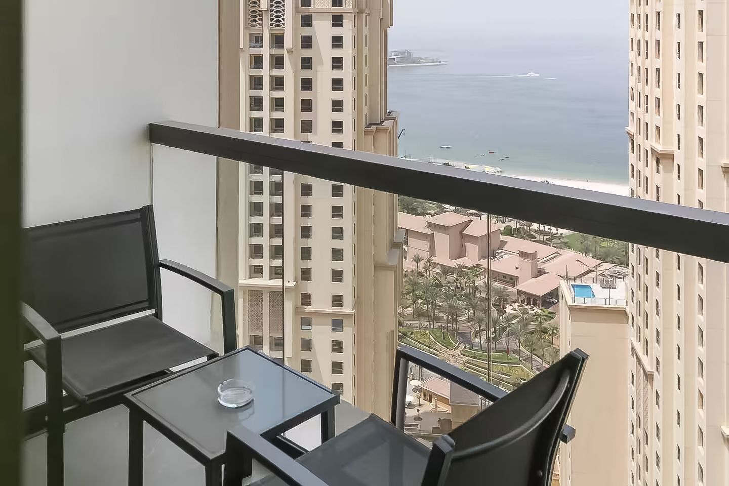 Een balkon met een glazen tafel en stoelen, met uitzicht op een hoog gebouw en een wateroppervlak.