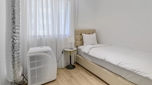 Ein kleiner, ordentlicher Raum mit einem Bett, einem Nachttisch mit Lampe, einem Heizlüfter und durchsichtigen Vorhängen.