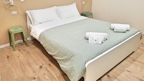 A neatly made bed with white linens, a green throw blanket, and a set of towels on the bed. There is a green nightstand with a lamp and a power outlet on the wall. - B B Palazzo Zappala (Catania)