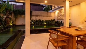 A modern outdoor dining area with a pool, wooden furniture, and decorative lighting. - Juada Garden (Seminyak)