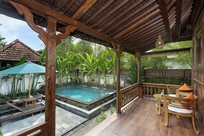 A covered outdoor area with a pool, wooden furniture, and a view of a house in the background. - Meng Bengil Villa (Tegallalang)