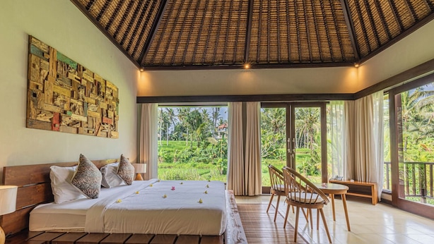 A bedroom with a large bed, a wooden headboard, a wall art piece, a window with a view of greenery, and a balcony with a chair.
