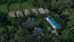 An aerial view of a resort with thatched-roof huts, a swimming pool, and lush tropical vegetation.
