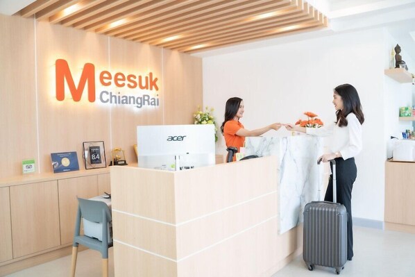 A reception desk with a sign reading "Meesuk ChiangRai" and an Acer computer. There is a blue chair and a grey suitcase. - Meesuk Residence (Chiang Rai)