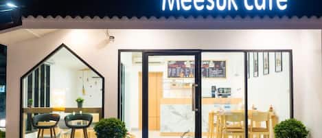 Una cafetería llamada 'Meesuk cafe' con un diseño moderno, que cuenta con un amplio escaparate de vidrio, un letrero acogedor y una zona de estar con sillas y mesas.