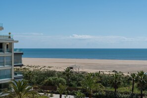 Blick auf einen Strand von einem modernen Gebäude mit Glasfassade, im Vordergrund Palmen und ein klarer, blauer Himmel.