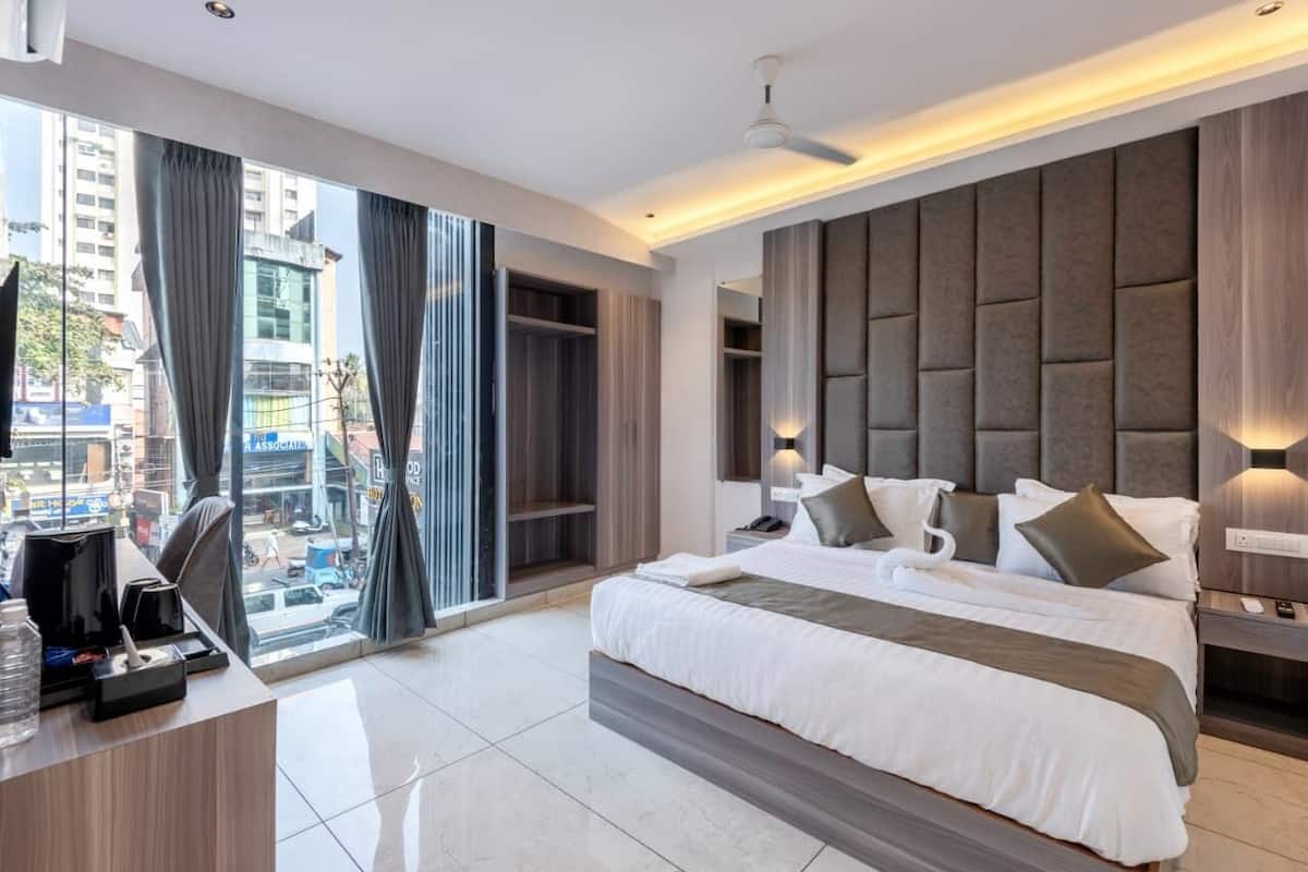 A modern hotel room with a large bed, a desk with a computer, and a view of the city through large windows.