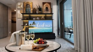 Camera d'albergo moderna con una grande TV che mostra una spiaggia, un cartello di benvenuto, una ciotola di frutta e una bottiglia di champagne.