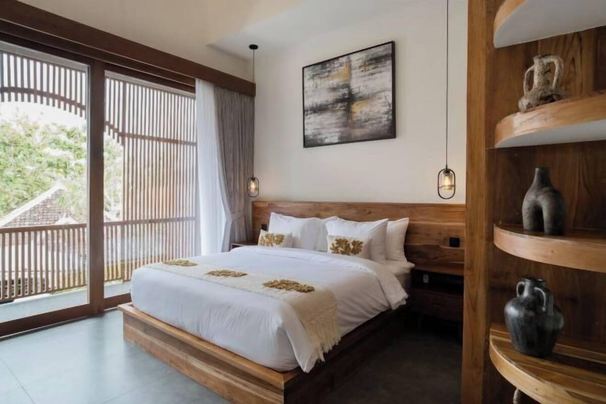 A bedroom with a wooden bed, a large window with a view, a wall-mounted abstract painting, and a shelf with decorative items.
