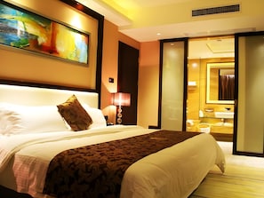 Hotelkamer met een groot bed, een schilderij aan de muur, een lamp en een zichtbare badkamer achter een glazen deur.