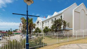 A white building with a red roof, a fenced area with palm trees, and a street lamp. - Road Lodge Gqeberha (Gqeberha)
