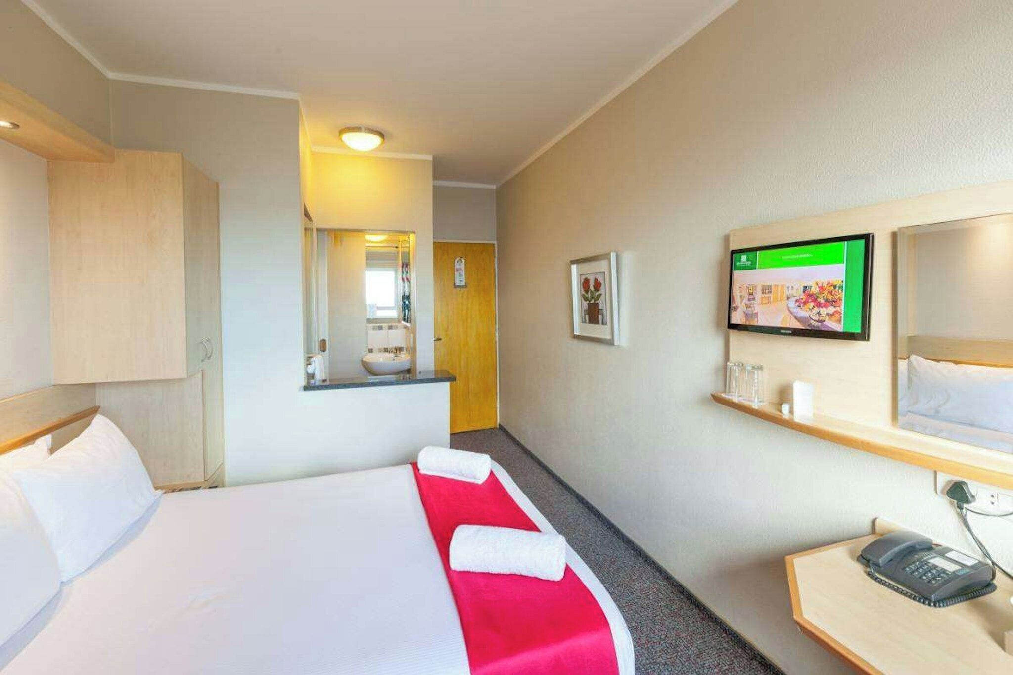Habitación de hotel con cama, escritorio con teléfono, televisor montado en la pared y un espejo.