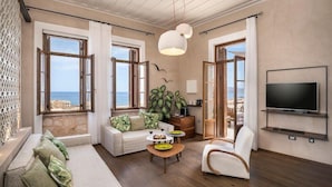 A spacious living room with a large TV, a sofa, a dining table, and a view of the sea through open windows.