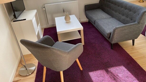 A modern living room with a grey sofa, a grey armchair, a white table, and a floor lamp. There are two windows with potted plants on the windowsill. - Herning City Apartments (Herning)