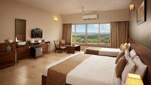 A hotel room with a large bed, a desk, a chair, a small fridge, a television, and a window with a view of the city. - At Home B&B Dona Paula (Panaji)