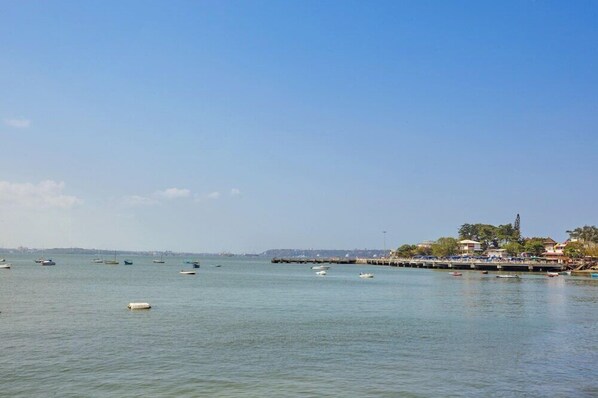 Vista costera con barcos en el agua, un muelle y edificios al fondo.