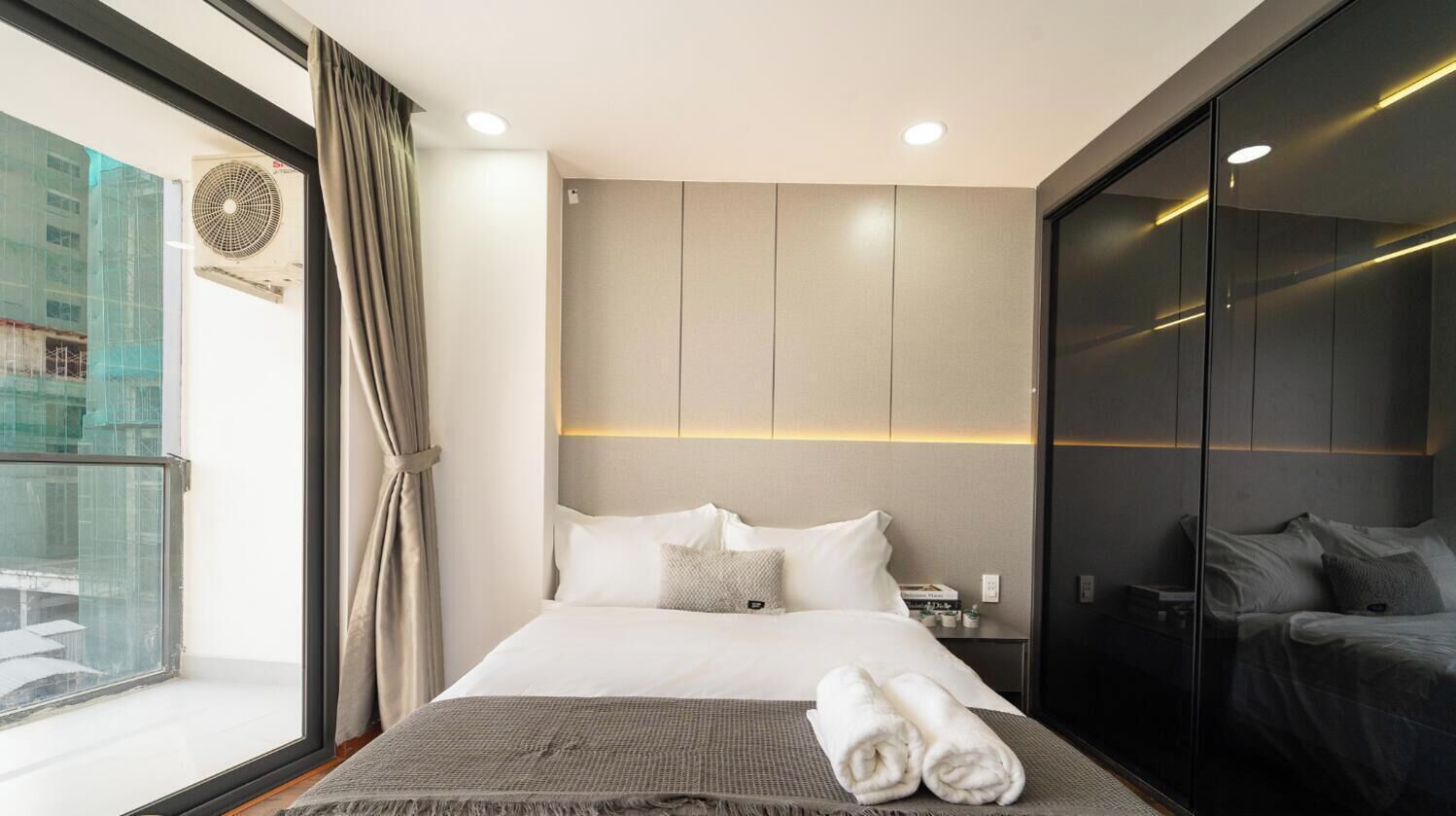 A modern hotel room with a large bed, a bedside table, a wardrobe, and a view of the city through a large window.