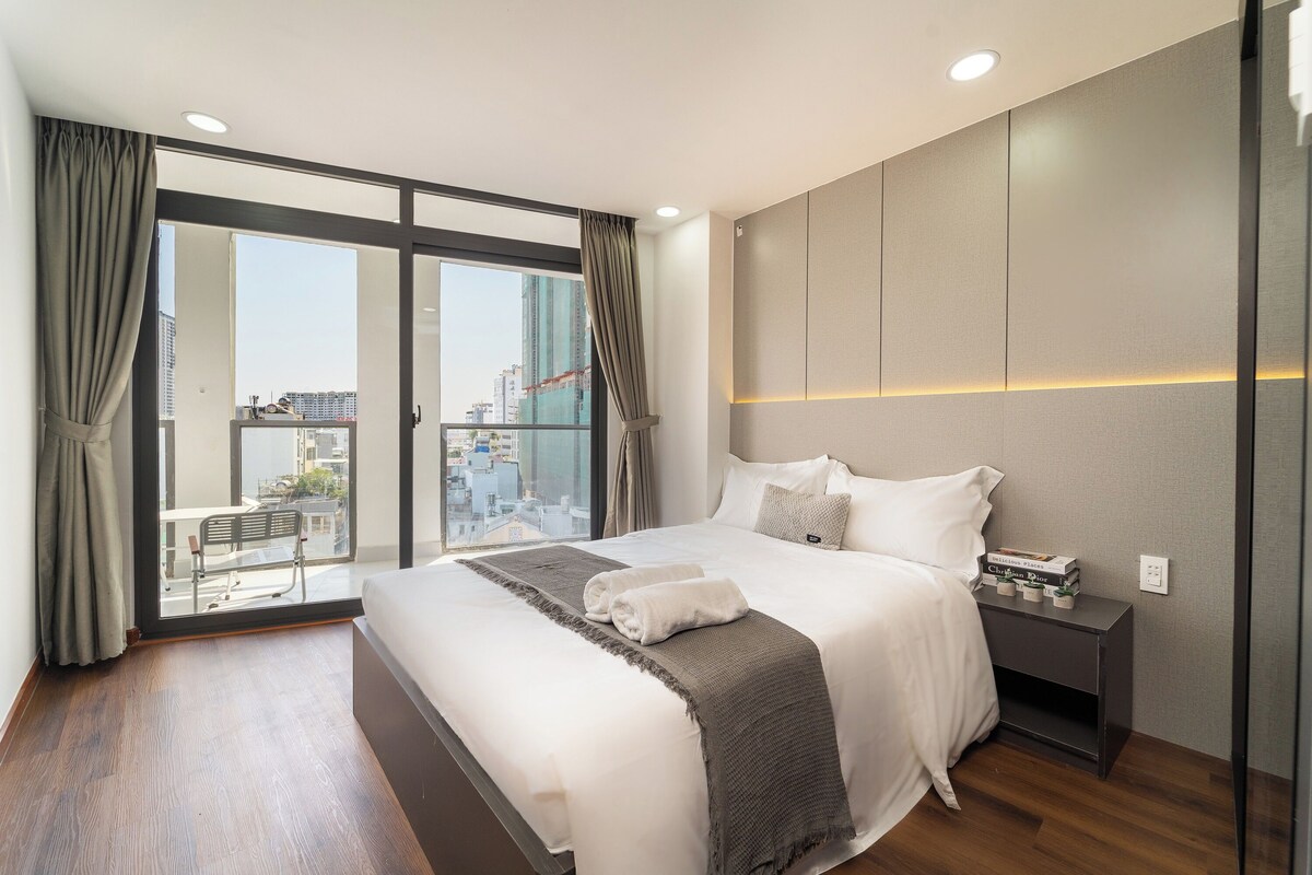 Ein modernes Hotelzimmer mit einem großen Bett, einem Nachttisch und Blick auf die Stadt durch raumhohe Fenster.