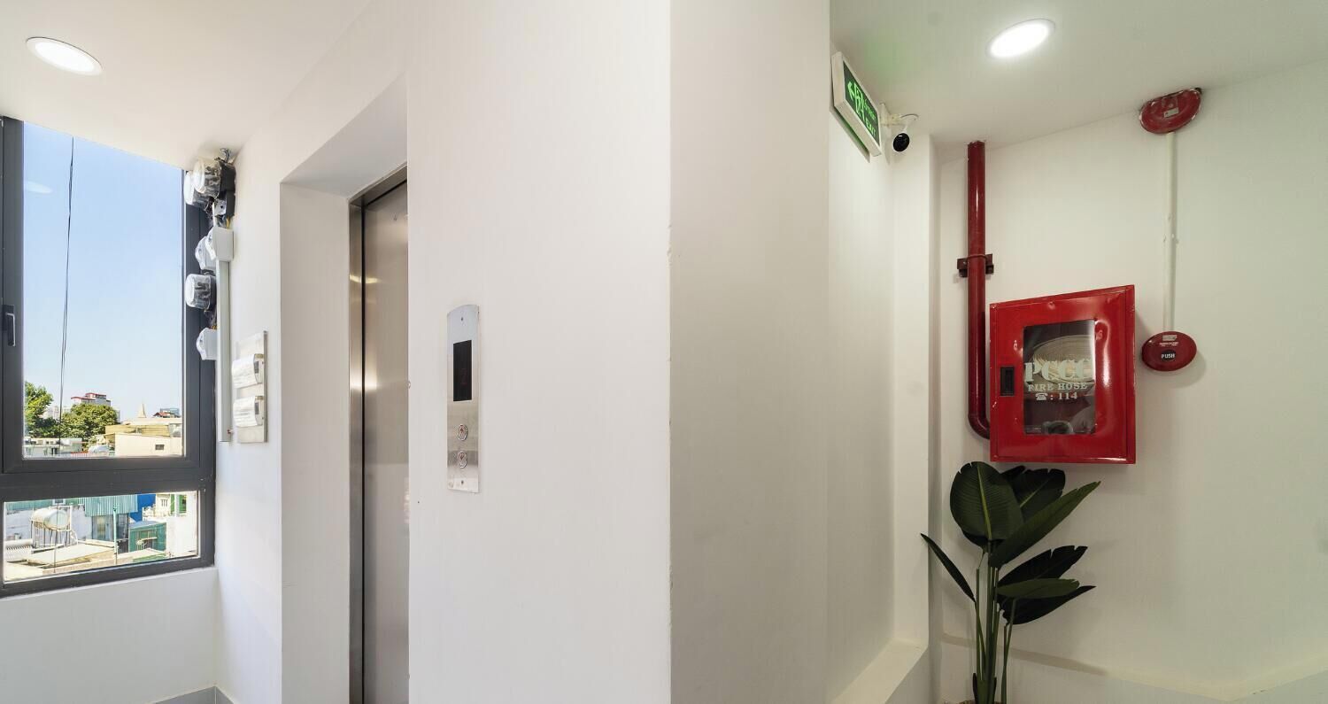 A hallway with a door, an elevator, a red emergency button, a security camera, and a plant.