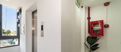 A hallway with a door, an elevator, a red emergency button, a security camera, and a plant.
