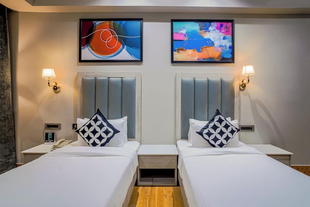 A hotel room with two beds, each with a headboard, a bedside table, a lamp, and a phone. There are two paintings on the wall above the beds.