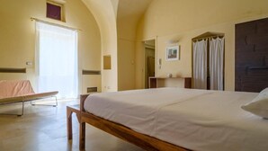 A bedroom with a large bed, a wooden bench, a small table, and a window with curtains. - Masseria Bernardini (Nardò)