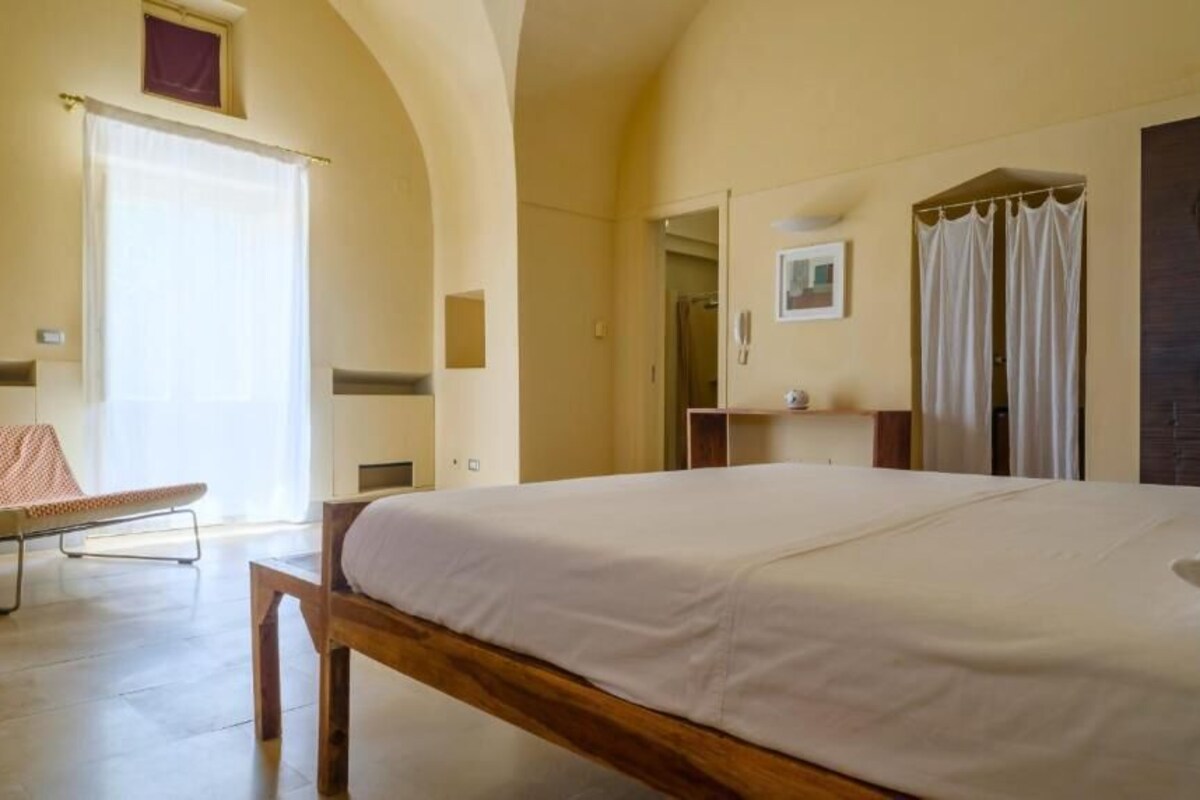 A bedroom with a large bed, a wooden bench, a small table, and a window with curtains.