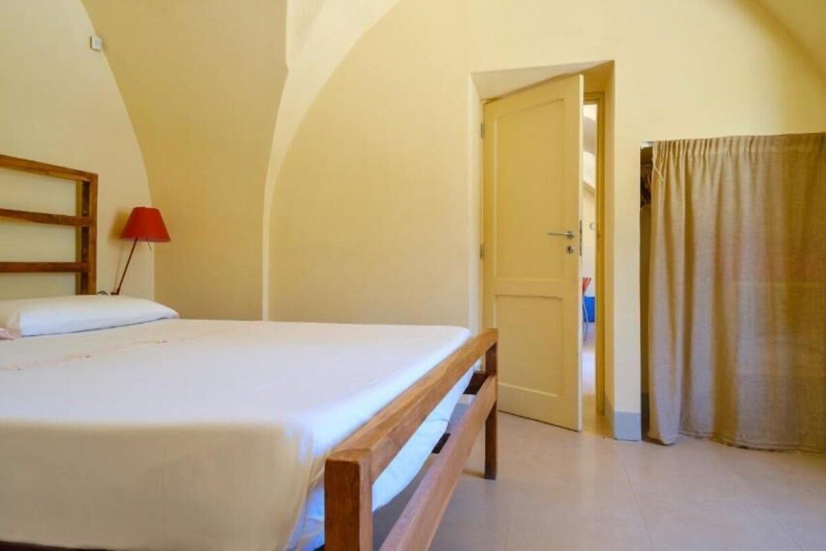 A bedroom with a bed, a wooden headboard, a red lamp, and a door leading to another room.