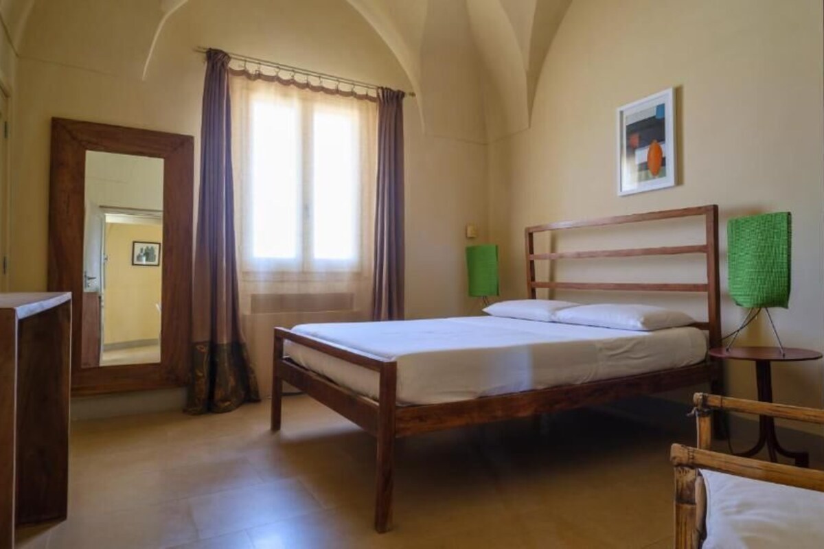 A bedroom with a wooden bed, a bedside table, a green lamp, and a painting on the wall.