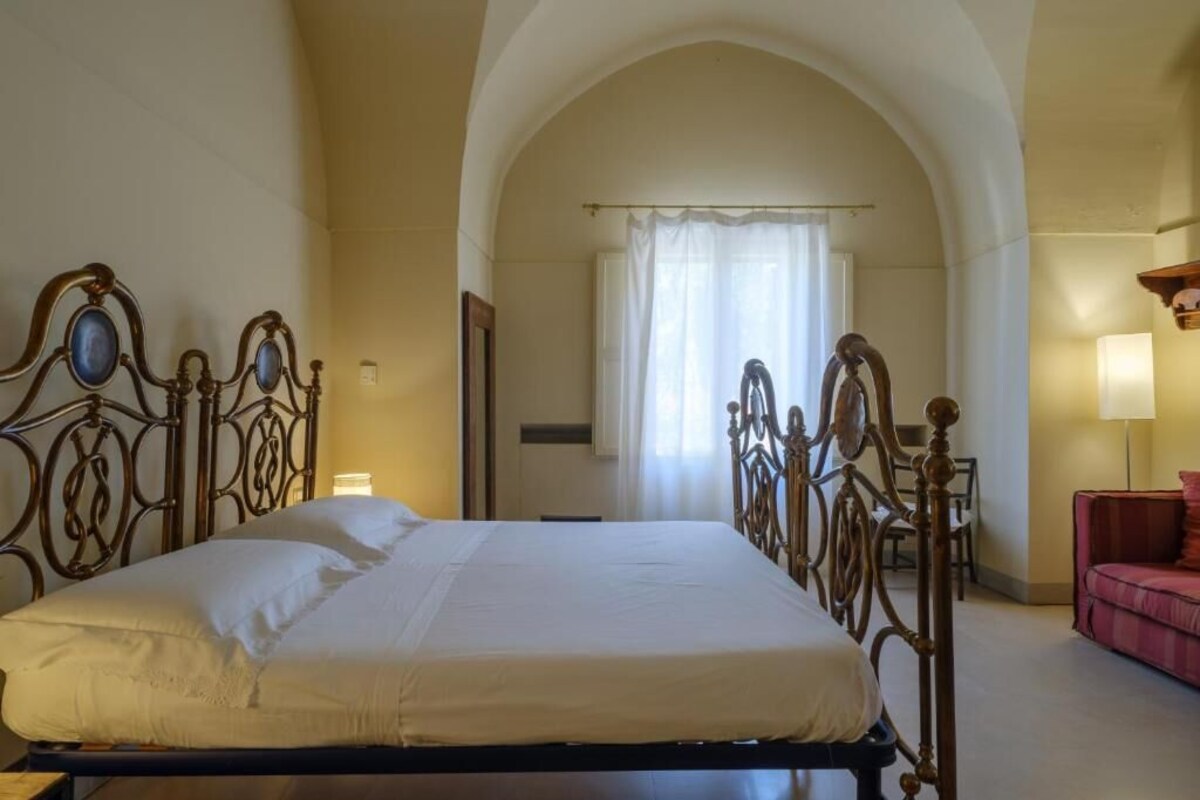 A neatly made bed in a room with an arched doorway, a small table with a lamp, and a red chair.