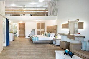 Una habitación de hotel moderna con cama de altillo, zona de estar con sofá, una mesita y un baño visible al fondo.