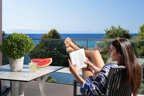 Una persona leyendo un libro en un balcón con vistas al mar, una planta en maceta y una rebanada de sandía sobre la mesa.