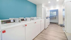 A laundry facility with multiple washing machines, a blue wall, and a window with blinds.